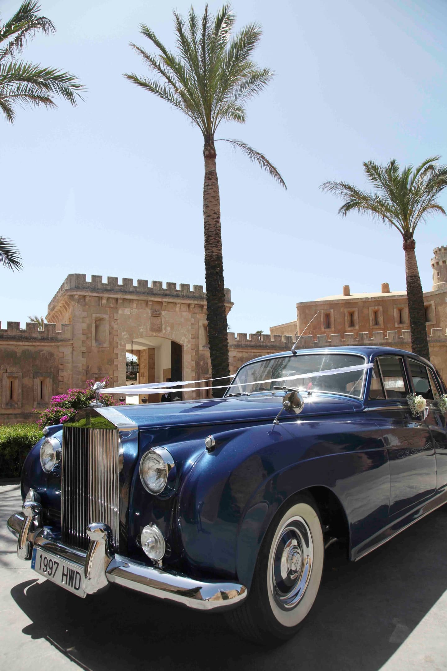 A classic navy Rolls-Royce parked in front of a stone castle in Mallorca under palm trees