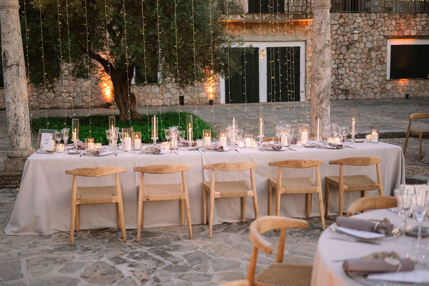 A long white dinner table with wooden chairs set in a walled courtyard