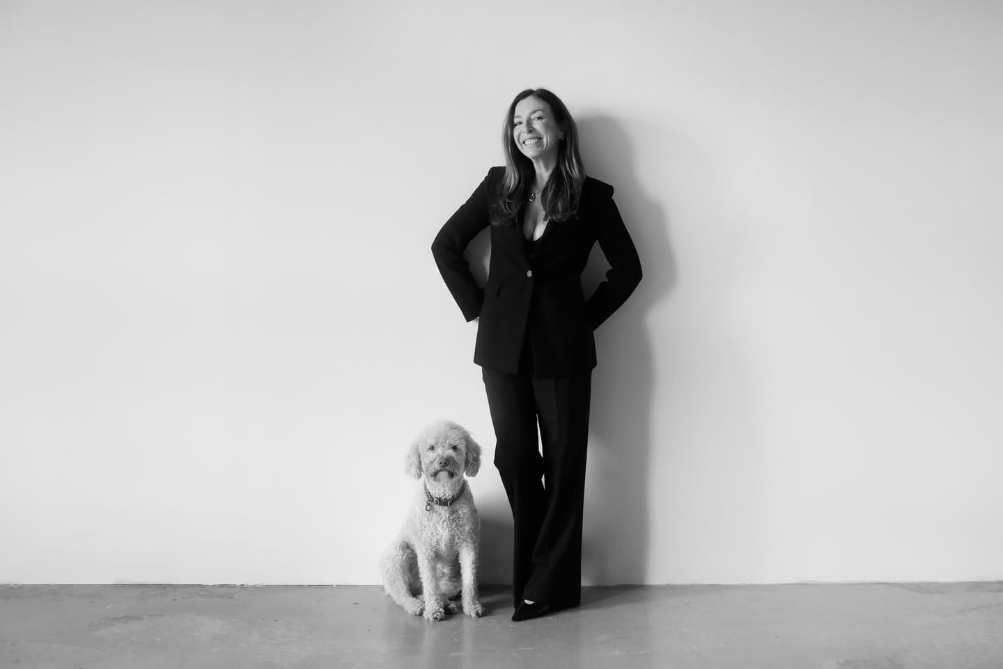 Luisa Catalán, founder of The Label & Co., in a black suit against a white wall with her poodle beside her