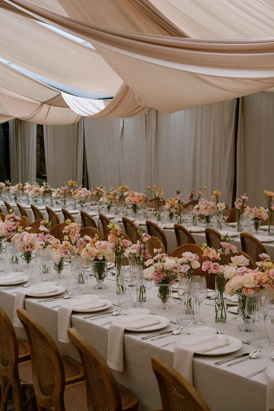 A long wedding table dressed with pastel flowers and glassware inside a draped marquee